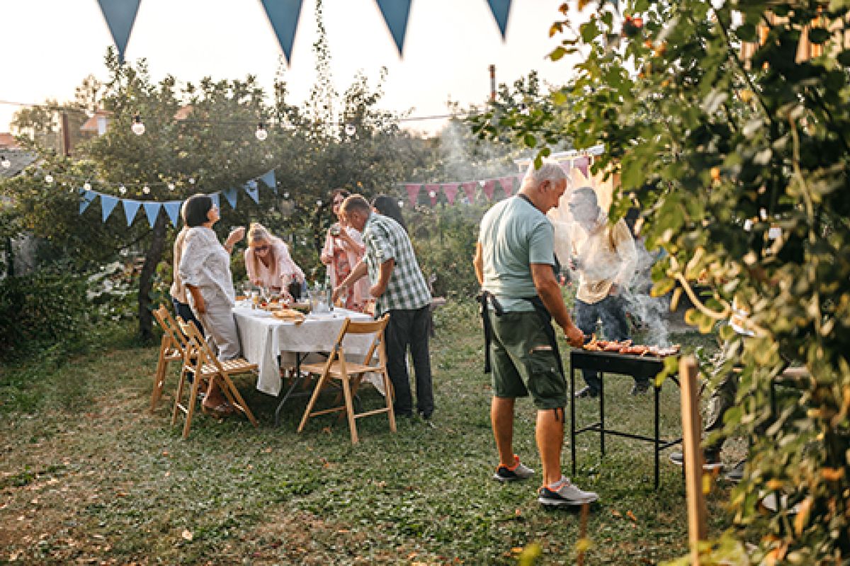 Mann grillt Fleisch im Garten, während die Familie den Tisch für das Mahl deckt.