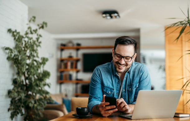 Porträt eines fröhlichen Mannes, der im Homeoffice ein Smartphone benutzt.
