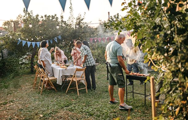 Mann grillt Fleisch im Garten, während die Familie den Tisch für das Mahl deckt.
