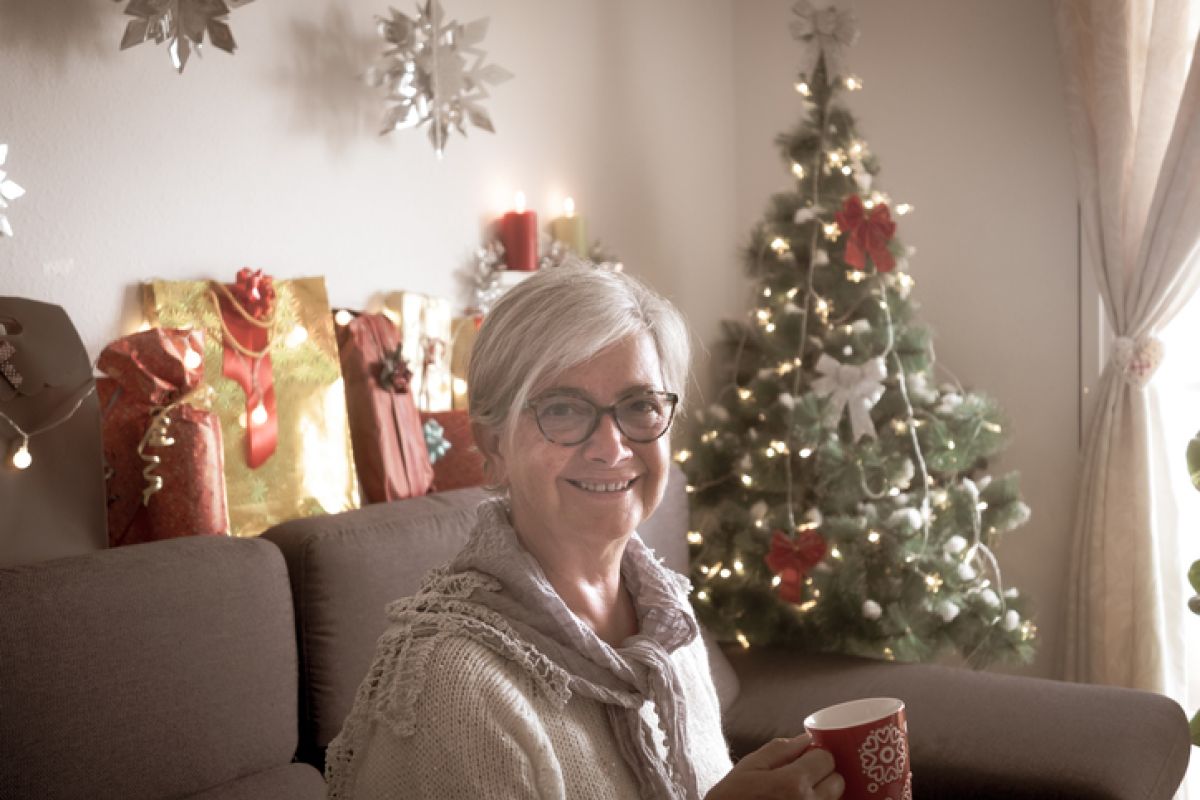 Glückliche Gloeckle Gewinnerin sitzt mit einer Tasse in der Hand auf der Couch, während im Hintergrund ein Weihnachtsbaum steht.