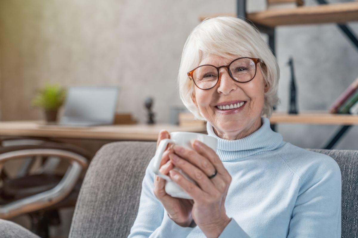 Eine ältere Frau sitzt auf der Couch mit einer Tasse in der Hand und ist eine SKL-Gewinnerin.