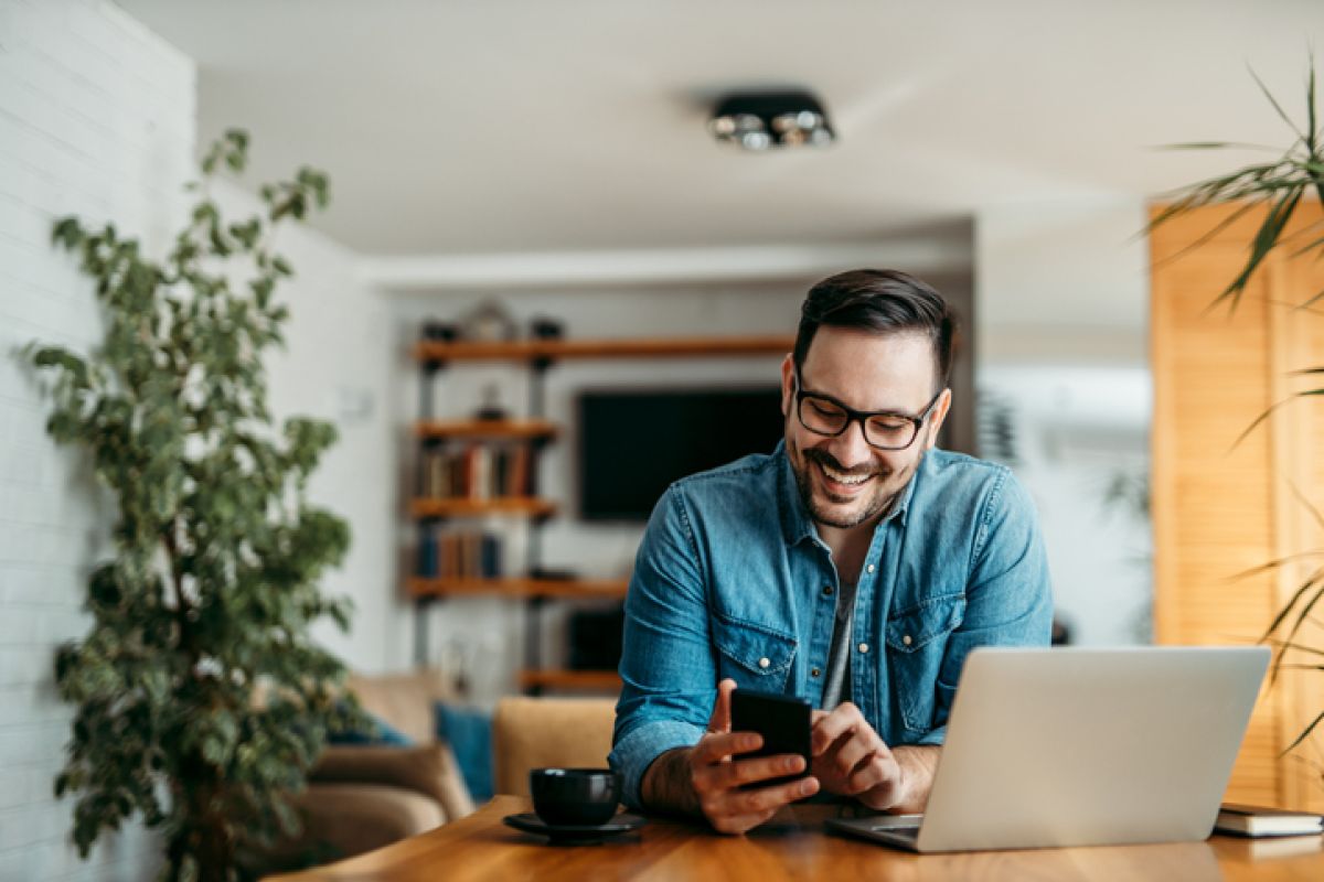 Porträt eines fröhlichen Mannes, der im Homeoffice ein Smartphone benutzt.