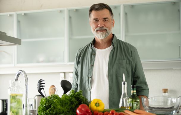 Lächelnder reifer Mann steht in der Küche, schaut in die Kamera und bereitet alleine zu Hause einen frischen Gemüsesalat zu.