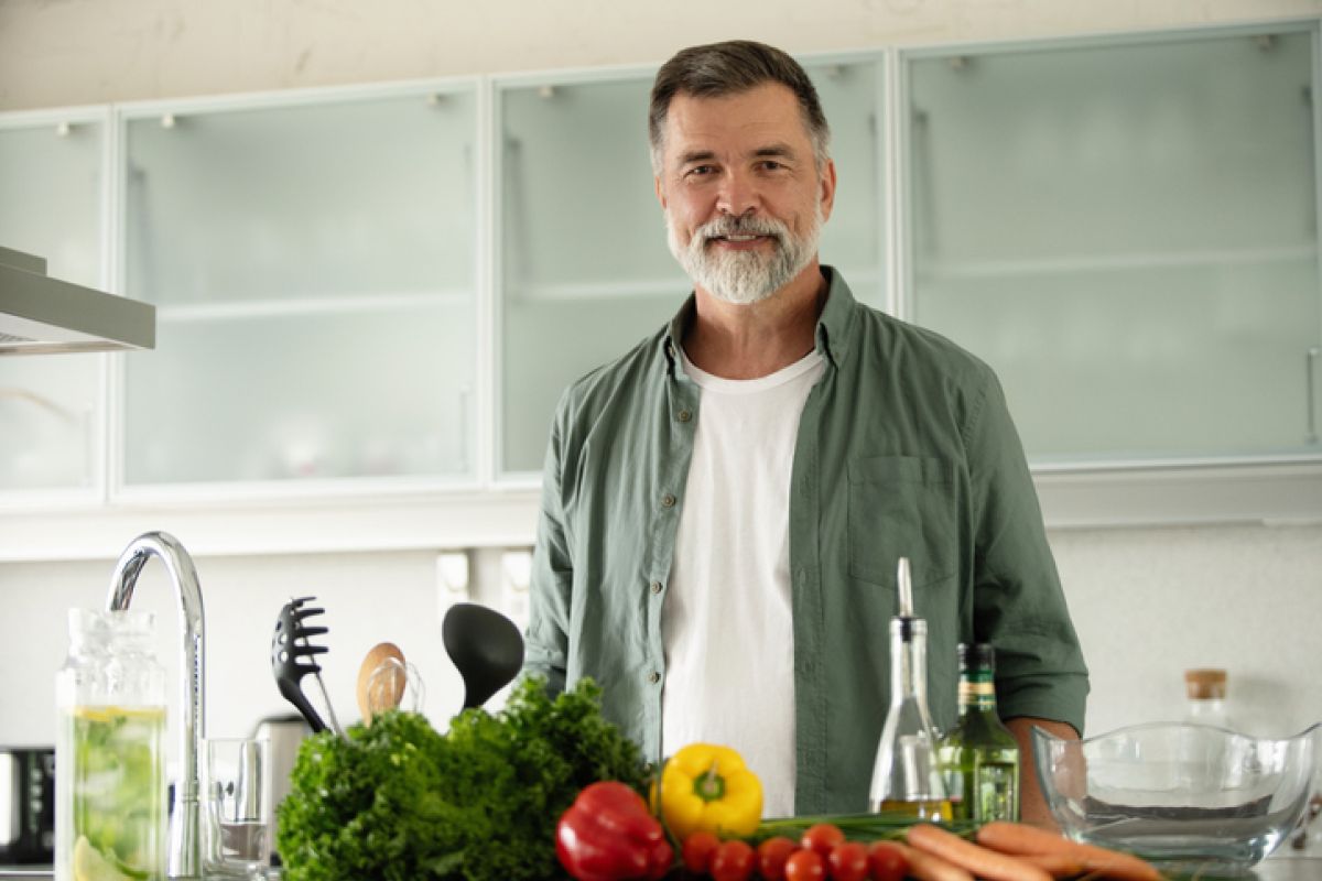 Lächelnder reifer Mann steht in der Küche, schaut in die Kamera und bereitet alleine zu Hause einen frischen Gemüsesalat zu.